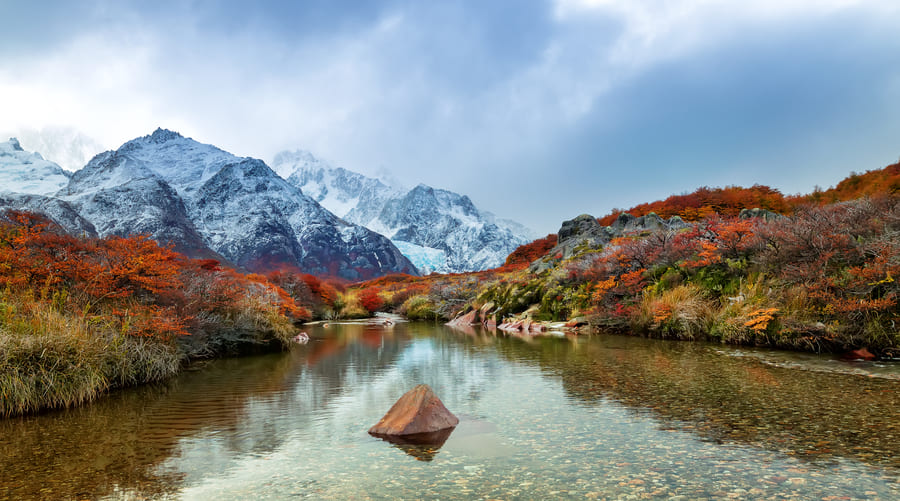 Un lago alpino rodeado de vegetación rojiza con cumbres nevadas.