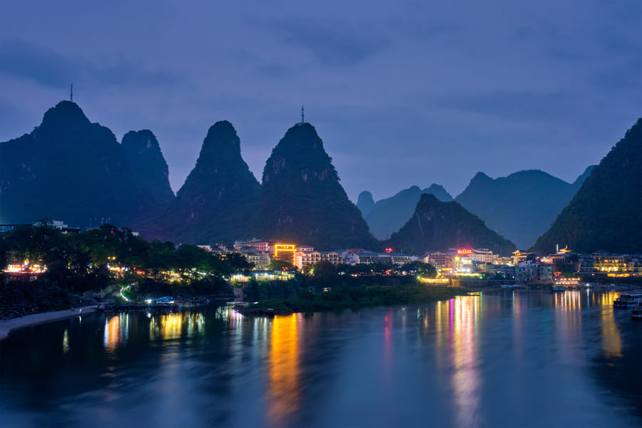 Un pueblo iluminado junto al río con montañas kársticas al anochecer.